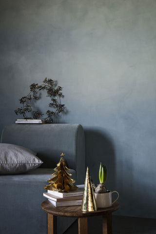 A styled interior scene with a blue-gray upholstered sofa against a textured blue-gray wall, a small round wooden side table with books and decorative gold tree ornaments, a gray pillow, and a potted plant.