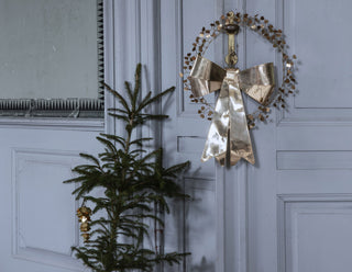 A metallic gold Christmas wreath with a large bow and small gold leaf accents, hanging on a light blue paneled door next to a small evergreen tree.