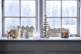 Decorative window sill with Christmas tree, candles, and lace decorations against a snowy window backdrop.