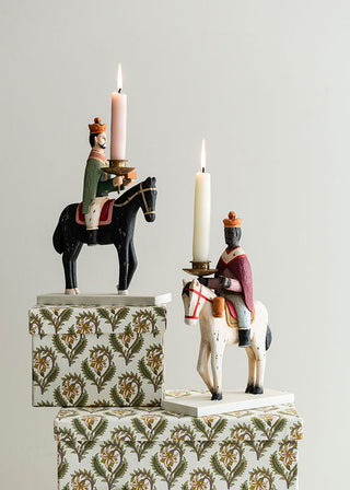 Decorative candle holders with figurines of men on horseback on patterned boxes against a gray background.