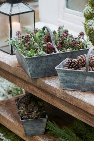 Metal baskets filled with frosted winter berries and pinecones on a snow-dusted wooden shelf beside a lit lantern