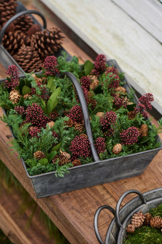 Overhead view of a metal basket filled with evergreen foliage, red berries, and pinecones next to a basket of pinecones