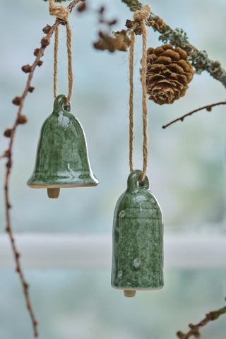 Two green ceramic bell ornaments hanging from a frosted branch beside a pine cone, against a soft blue-toned background