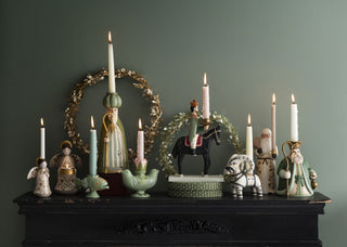 A decorative holiday mantel display featuring multiple ceramic candle holders, lit taper candles, a green wreath, and figurines including Santa Claus, an angel, a horse rider, and a white dog, set against a dark green wall.