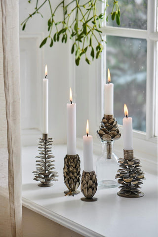 Five metal pinecone candle holders of varying heights and finishes holding lit white candles on a white windowsill, with mistletoe hanging above and a glass bottle among them