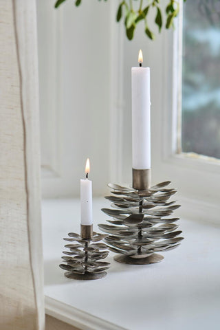 Two pine cone-shaped candle holders in different sizes holding lit white taper candles on a white windowsill