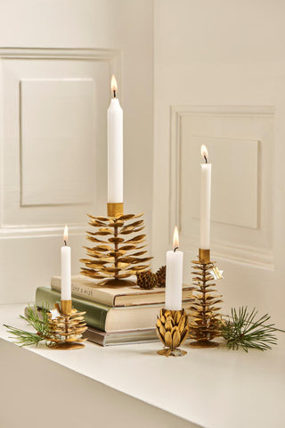 Four brass pinecone candle holders in different heights with lit white taper candles, styled on stacked books with pinecones and pine sprigs