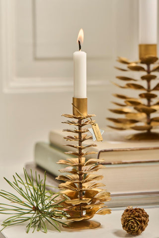 Close-up of a lit candle in a brass pinecone candle holder with a gift tag, a pine sprig, and a small pinecone on a shelf, another holder blurred behind