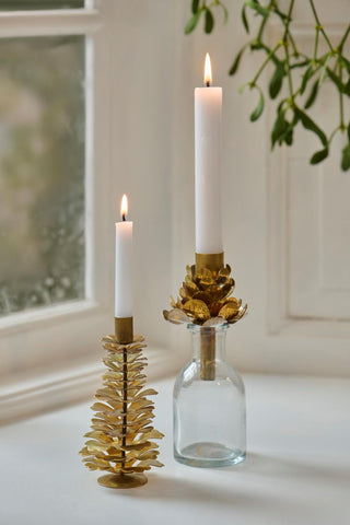 Two brass pinecone candle holders with lit candles by a window, one set into a small glass bottle, with mistletoe hanging above