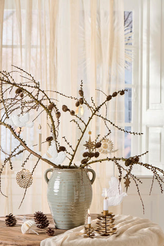 Two brass pinecone candle holders with lit white candles on a linen cloth beside a ceramic vase with festive branch decorations