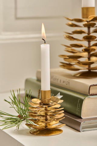Small brass pinecone candle holder with a lit candle and pine sprig, set on a stack of books beside a taller matching candle holder