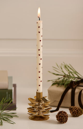 Lit taper candle with a red heart pattern in a brass pinecone candle holder, beside books and a wrapped gift