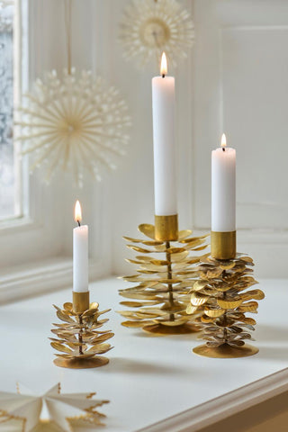 Three brass pinecone candle holders of different heights with lit candles on a windowsill with paper star decorations