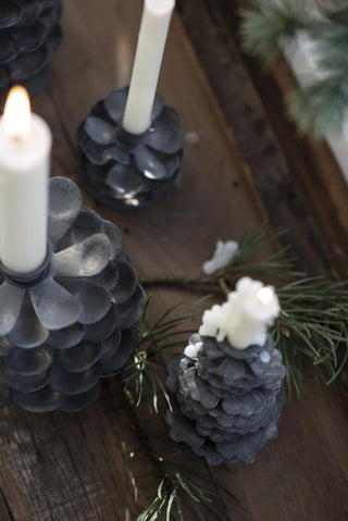 Grey metal pinecone candle holders of different sizes on a dark wood table with lit and unlit white candles and pine sprigs