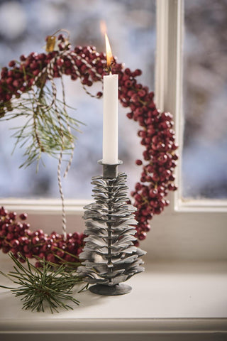 Close-up of a small grey metal pinecone candle holder with a lit white candle on a windowsill next to a red berry wreath
