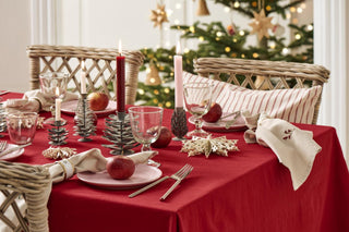 Christmas table setting with a red tablecloth, grey metal tree- and pinecone-shaped candle holders holding lit candles, apples, wicker chairs, and a decorated Christmas tree in the background