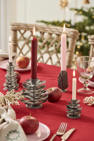 Grey metal tree- and pinecone-shaped candle holders with lit candles, red and pink candles, apples, and paper stars on a red Christmas table setting