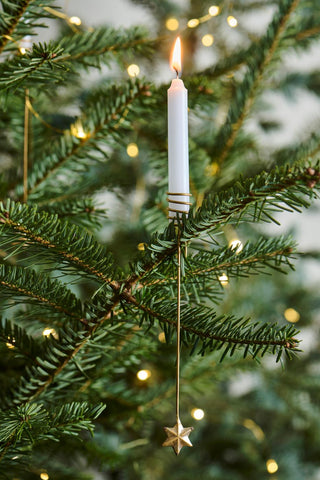 Gold brass candle clip holding a lit white taper candle on a Christmas tree branch, with a small star hanging below and string lights in the background