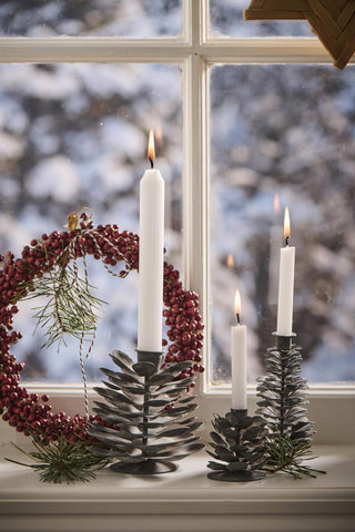 Grey metal pinecone-shaped candle holders in three sizes with lit candles on a windowsill beside a red berry wreath, snowy view outside