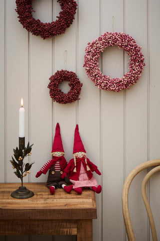 Metal leaf and wooden bead candle holder with a lit candle on a wooden table beside two gnome dolls and red and pink berry wreaths hanging on the wall