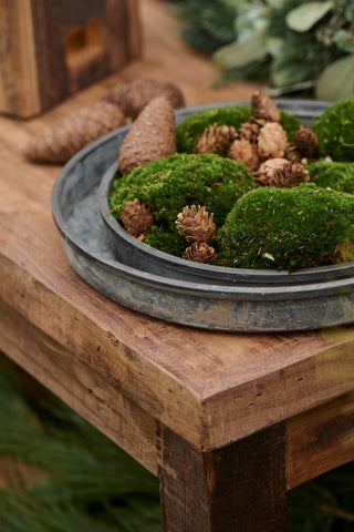 Round weathered metal tray styled with moss and pinecones as a Christmas table centrepiece on a rustic wooden table