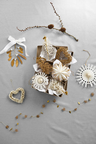 Assorted white and gold beaded and paper Christmas ornaments including trees, stars, a heart, and a mistletoe sprig, styled in a gift box beside a pinecone branch