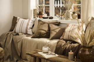 Chocolate brown velvet cushion cover among striped linen and beige pillows on a daybed by a window, with a coffee cup, open book, and ceramic vases nearby