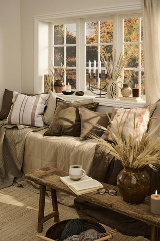 Wide shot of a cosy window nook with striped and chocolate brown velvet cushions, a brass candelabra, dried wheat in vases, and a wooden bench with coffee and a book
