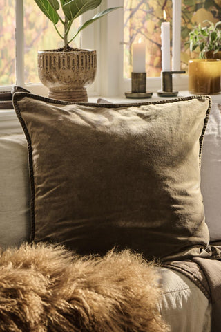Close-up of a chocolate brown velvet cushion propped against a striped pillow near a potted plant, lit candles, and a sheepskin throw