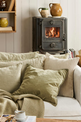 Green velvet cushion among other cushions and a wool throw on a light sofa in front of a lit wood-burning stove