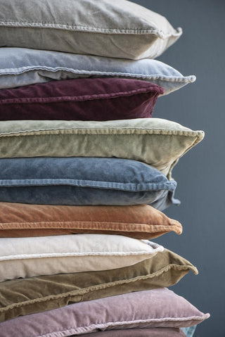 Stack of stonewashed cotton velvet cushions in assorted colours including grey, blue, burgundy, sage green, and slate, showing the piped-edge finish