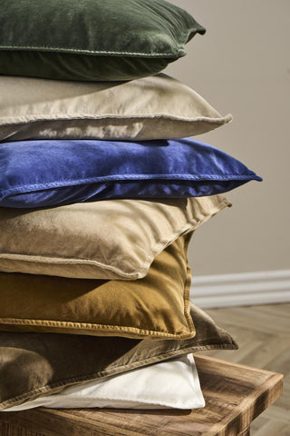 Stack of velvet cushions in olive green, taupe, cobalt blue, and mustard tones on a wooden table