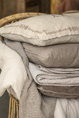 Stack of grey, taupe and cream velvet and linen cushions with a striped quilted throw and an embroidered linen cushion on a wicker chair