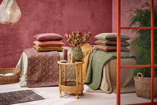 Stacks of velvet cushions in pink, terracotta, ochre, and green tones on quilted throws and a floral bedspread against a rose-coloured textured wall