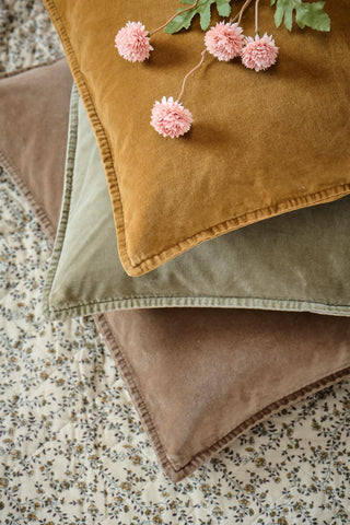 Close-up of stacked ochre, sage green, and brown velvet cushions with stitched piping, topped with dried pink flowers on a floral quilted throw