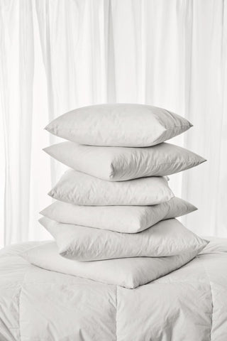 A neat stack of six white cushion fillers on a folded white quilt against a sheer white curtain