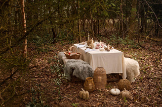 Woodland autumn table set with fur throws, a wicker basket, rattan lanterns, and seagrass and fabric pumpkins on the ground