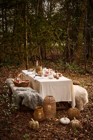 Autumn forest table setting with candlelight, a basket of apples, fur throws, and woven seagrass pumpkins beside rattan lanterns