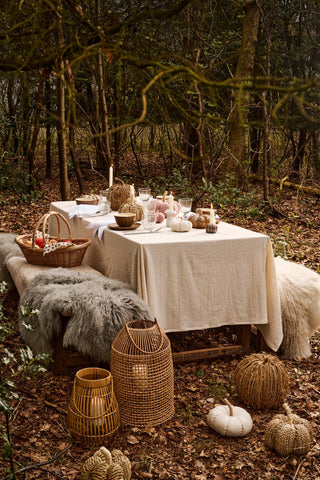 Rattan lanterns, a woven basket, and seagrass and fabric pumpkins on fallen leaves in front of a forest dining table