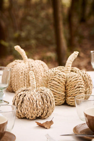 Close-up of woven seagrass pumpkins of different sizes with coiled stems, arranged on a table beside glassware and a bowl