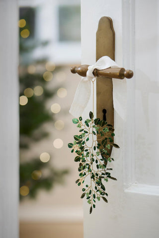 Green beaded leaf ornament hanging from a vintage brass door handle wrapped with a white ribbon, with a lit Christmas tree visible through the open door