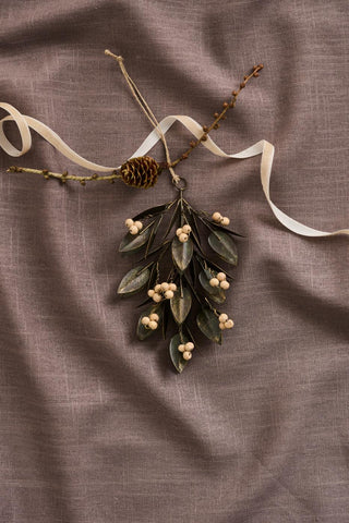 Metal leaf and wooden bead hanging ornament resting on a mauve linen cloth, next to a pine cone and sprigs of dried berries tied with a cream ribbon