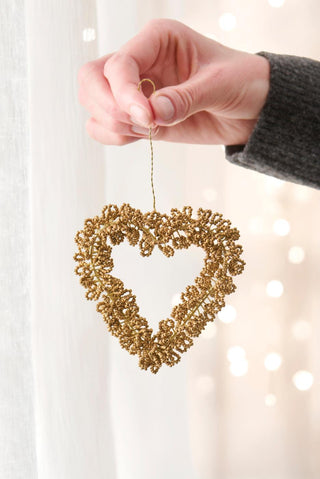 A hand holding a gold-brown beaded heart ornament by its wire loop, with soft fairy lights in the background