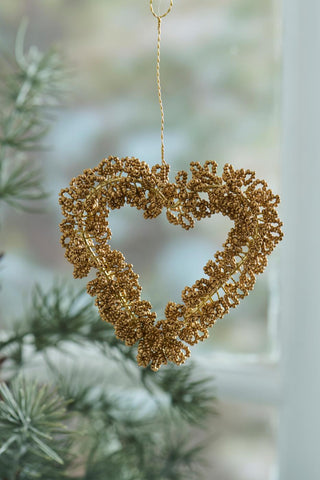 Gold-brown beaded heart ornament hanging in front of a pine branch and a window