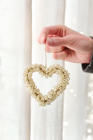 Hand holding a white beaded heart ornament by its gold cord in front of sheer white curtains