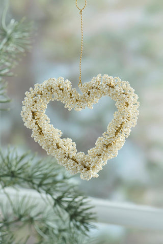 White beaded heart ornament hanging in front of a blurred pine branch and window