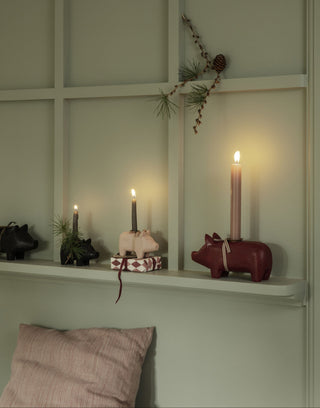 A styled interior shelf scene with candles and small decorative pig figurines, including a red pig holder with a tall lit candle and a pink pig figurine with a wrapped gift on top. A pale green paneled wall and shelf are visible, along with part of a pink pillow at the bottom.