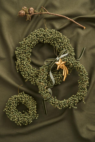 Gold shooting star ornament tied with a sage green ribbon to a wreath made of dried green berries, beside a small pinecone-tipped branch