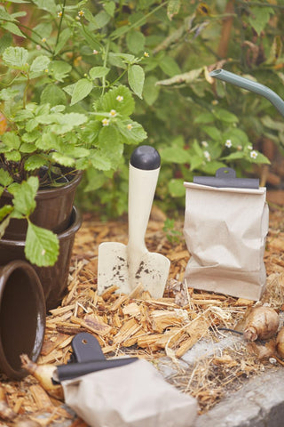 Kraft paper bags with clip closures beside a garden trowel and flower bulbs among garden plants