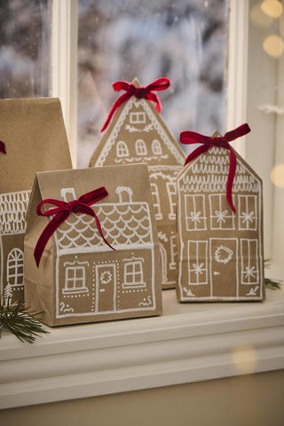 Kraft paper gift bags hand-painted as gingerbread houses with red ribbon bows on a snowy windowsill